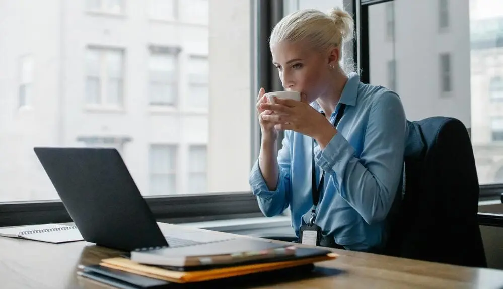 Indecisive young woman looking at laptop drinks coffee
